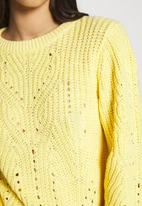 Pull en tricot jaune avec un motif Texturé, présentant des mailles aérées et un col côtelé. Le tissu semble doux et léger.