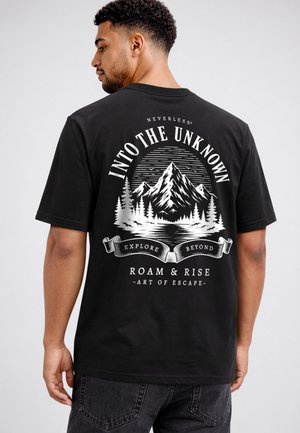 Mann trägt ein schwarzes T-Shirt mit weißem Berg- und Waldgrafik und dem Text "Into the Unknown, Explore Beyond, Roam & Rise, Art of Escape" auf dem Rücken.