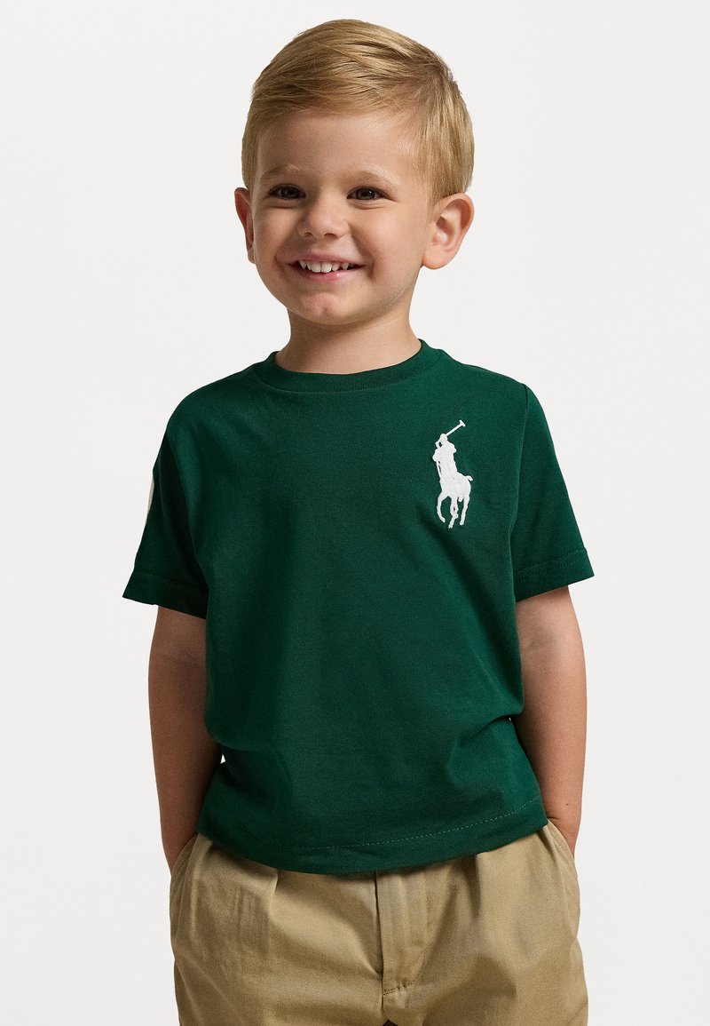 Grön bomullströja med kort ärm, med en vit polo-spelare logotyp på vänster bröst. Matchad med beige byxor.