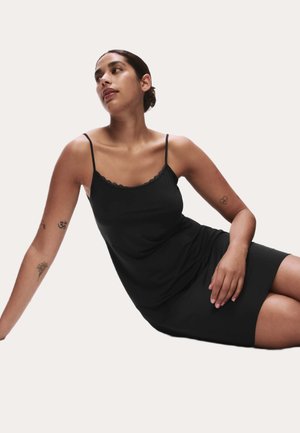 Kvinde med mørkt hår i en lav knold, iført en sort ærmeløs kjole, siddende med den ene arm strakt ud og kiggende til siden.