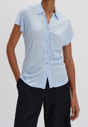 Osoba ubrana w jasno niebieską, krótkorękawną koszulę na guziki, luźno wsuniętą w czarne spodnie, stojąca z jedną ręką w kieszeni na jednolitym tle.