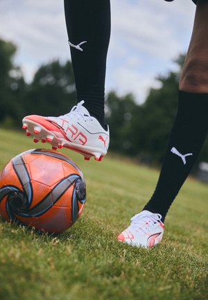 Hvite og rosa fotballsko med en teksturert såle, på gress, ved siden av en livlig oransje og blå mønstret fotball. Svarte sokker med logo.