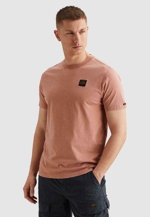 Junger Mann trägt ein ausgewaschenes rosa kurzärmeliges T-Shirt und schwarze Cargoshorts, steht mit einer Hand in der Tasche und schaut zur Seite.