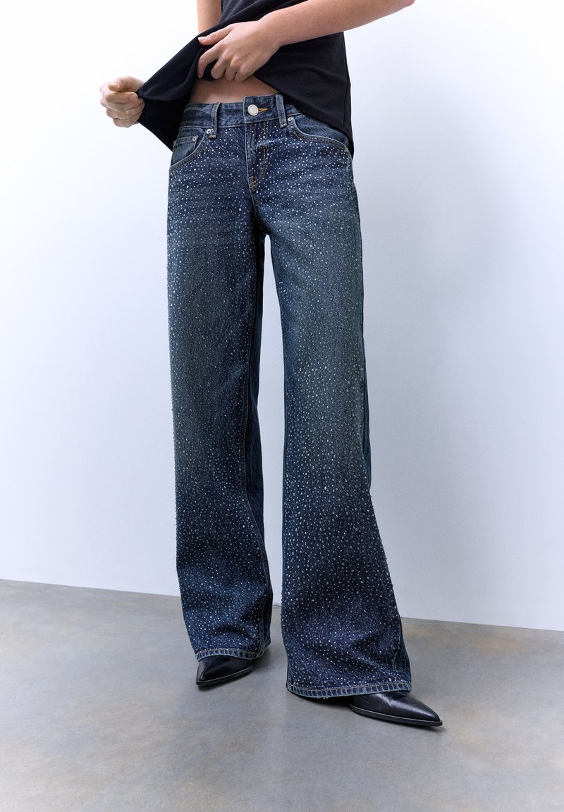 Leveälahkeiset tummansiniset farkut, joissa on siellä täällä strassikoristeita ja perinteinen viiden taskun malli, yhdistettynä kärjistetyihin mustiin kenkiin.