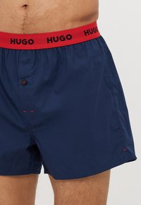 Boxers bleu marine avec une ceinture élastique rouge portant le logo "HUGO", fermeture à un bouton et détails de couture rouge contrastants.
