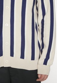 Beiger Cardigan mit navyblauen und weißen vertikalen Streifen, mit Knopfverschluss, gerippten Bündchen und Saum. Weiche, gestrickte Stofftextur.