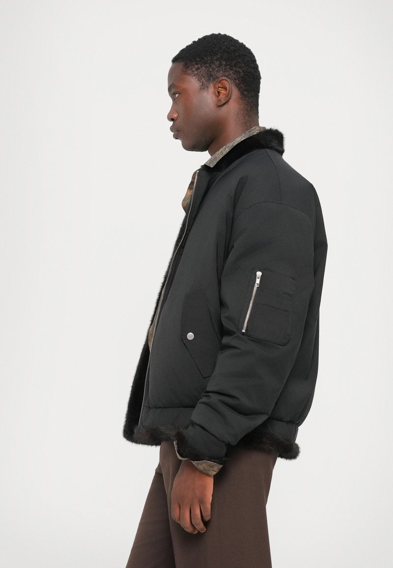Giubbotto bomber nero con pelo, tasca con zip sulla manica e finitura liscia. Caratterizzato da una silhouette aderente e polsini a costine.