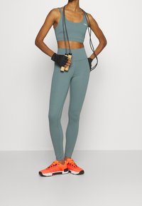 Smaragdowy stanik sportowy i legginsy z wysokim stanem w połączeniu z jasnopomarańczowym obuwiem sportowym; trzyma skakankę, ma na sobie czarne rękawice.