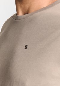 Lichtbruine t-shirt van een soepele stof met een ronde halslijn en een klein zwart lijnontwerp bij de kraag. Casual pasvorm, minimalistische stijl.