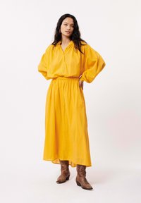 Robe chemise jaune à manches longues avec un devant boutonné et une taille élastique. Le mannequin porte des bottes de cow-boy marron, ajoutant une texture contrastante.