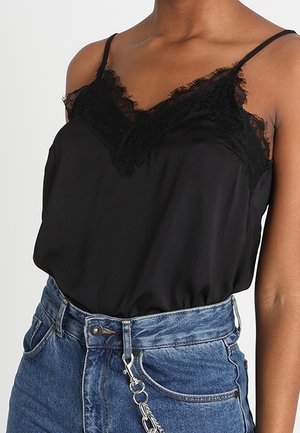Vrouw draagt een zwarte camisole met kanten rand, ingestopt in een high-waist blauwe spijkerbroek met een zilveren kettingaccessoire.