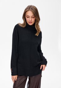 Kobieta z jasnobrązowymi włosami, ubrana w czarny sweter z golfem i ciemnobrązowe skórzane spodnie, patrzy w dół, z jedną ręką w kieszeni.