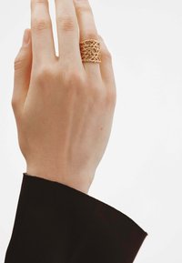 Anillo dorado con un intrincado diseño de entramado trenzado. Colocado en una mano con un tono de piel claro, que lleva una manga negra.
