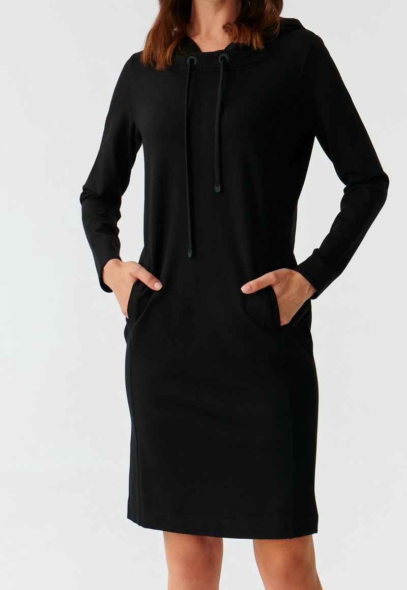Femme portant une robe à capuche noire à manches longues avec cordons, mains dans les poches avant, debout devant un fond uni.