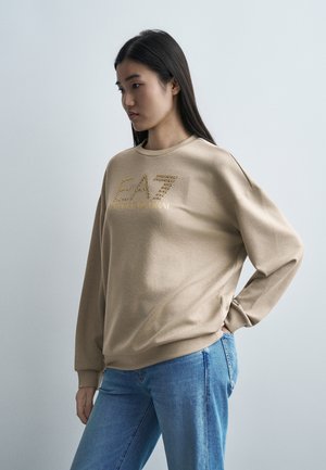 Giovane donna con lunghi capelli scuri che indossa una felpa beige EA7 Emporio Armani e jeans blu, in piedi contro uno sfondo chiaro e semplice.