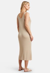 Knielanges, ärmelloses, beiges Kleid aus geripptem, strukturiertem Stoff mit tiefem Rückenausschnitt. Getragen mit flachen, beigen Sandalen.