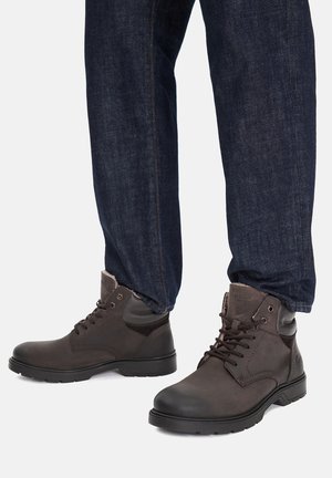 Legs wearing dark blue jeans and dark brown lace-up ankle boots with rubber soles on a plain light background.