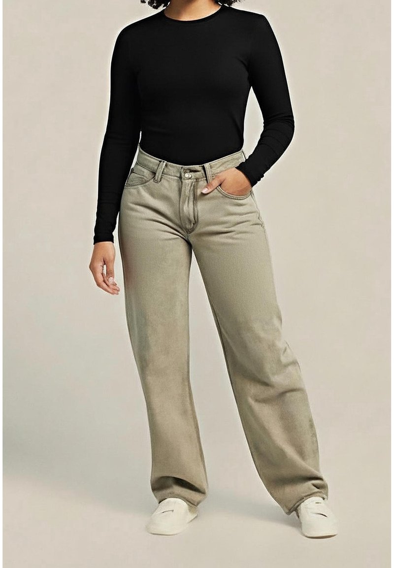 Personne portant un haut à manches longues noir, un pantalon beige ample, des baskets blanches, debout avec une main dans une poche sur un fond uni.