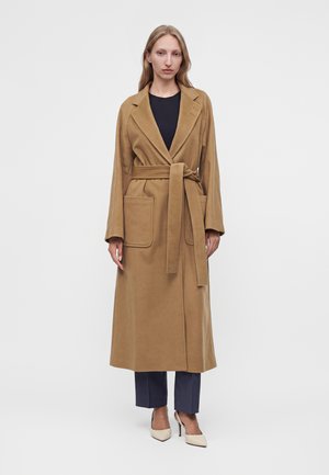 Mujer con un abrigo largo camel con cinturón, grandes bolsillos, pantalones azul marino y tacones blancos, de pie frente a un fondo blanco liso.
