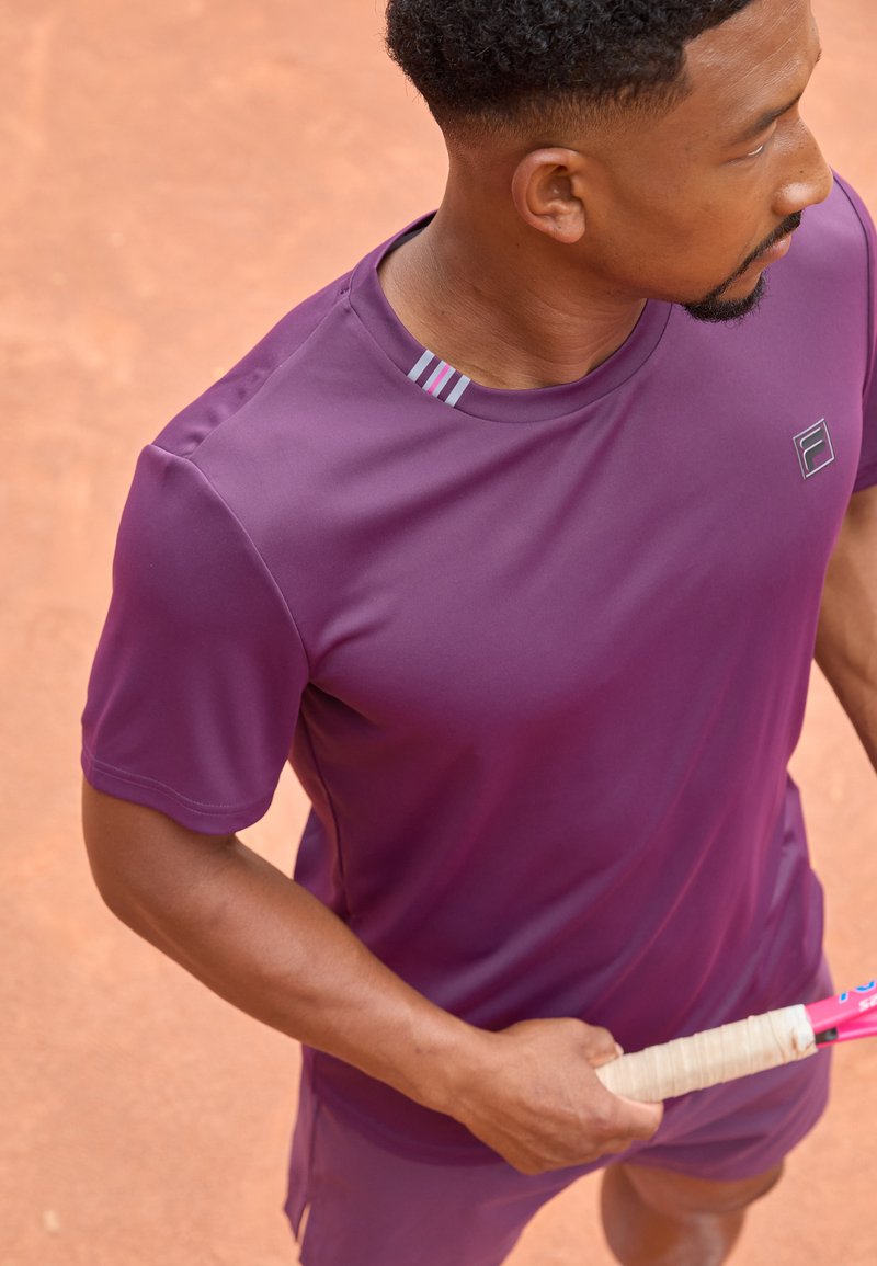 T-shirt de sport violet à manches courtes, avec un accent rayé sur le col. Le tissu est léger, adapté aux activités sportives.