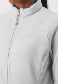 Giacca in fleece grigio chiaro con colletto alto, chiusura lampo completa e branding discreto sul petto. Texture morbida, senza motivi né accenti.