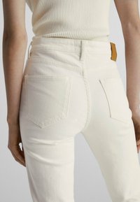 Jeans en denim blanc avec une taille haute, dotés de deux poches arrière et d'une étiquette en cuir beige au niveau de la taille, associés à un haut c ôtelé.
