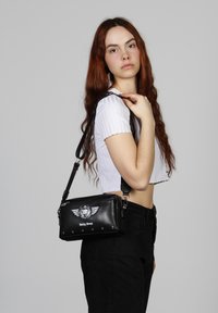 Bolso bandolera sintético negro con logo alado y tachuelas plateadas. Cuenta con una correa ajustable y cierre de cremallera. Diseño compacto rectangular.