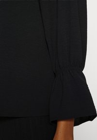 Blouse noire avec un tissu texturé, des manches bouffantes froncées et un poignet ajusté. Le design présente une finition lisse mettant en valeur une élégante simplicité.