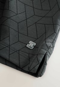 Svart tyg med geometriskt mönster och grå accenter, med en silverlogotyp och sydda kanter, som visar en mjuk textur.