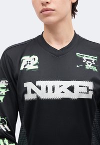Άτομο που φοράει μαύρο πουκάμισο Nike από δίχτυ με λευκά και πράσινα γραφικά λογότυπα και κείμενο, στέκεται μπροστά σε απλό ανοιχτό φόντο.