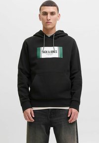 Junger Mann in schwarzem Hoodie mit "JACK & JONES"-Logo, kombiniert mit ausgebleichten schwarzen Jeans, steht vor einem schlichten Hintergrund.
