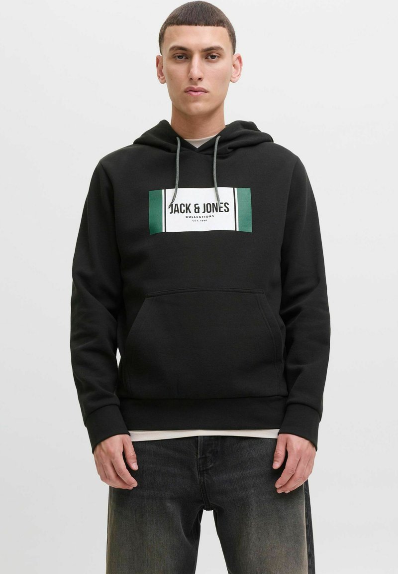 Junger Mann in schwarzem Hoodie mit "JACK & JONES"-Logo, kombiniert mit ausgebleichten schwarzen Jeans, steht vor einem schlichten Hintergrund.