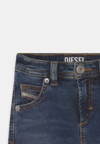 Jeans de denim azul escuro com costura laranja, design de cinco bolsos e fecho em botão prateado. Etiqueta "DIESEL" visível na parte interna da cintura.