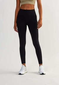 Leggings noires taille haute avec une texture lisse et des poches latérales, associés à des chaussures de sport blanches. Présentés sur un fond neutre.