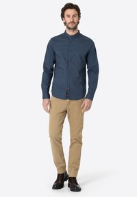 Navyblaues Hemd mit Stehkragen, strukturierter Stoff und kontrastierenden Knöpfen; kombiniert mit khakifarbenen Hosen und dunkelbraunen Stiefeln.