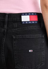 Melna džinsa bikses ar redzamu aizmugurējo kabatu, uz kuras ir zila, sarkana un balta austs etiķete ar uzrakstu "TOMMY JEANS". Tekstūrēta auduma.