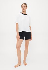 Frau, die barfuß steht und ein weißes, übergroßes T-Shirt mit schwarzem Kragen und schwarze Shorts trägt, vor einem schlichten weißen Hintergrund.