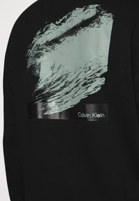 Felpa nera con una grafica a onda verde menta texturizzata e un'etichetta rettangolare nera lucida che presenta la scritta "Calvin Klein" in lettere bianche.
