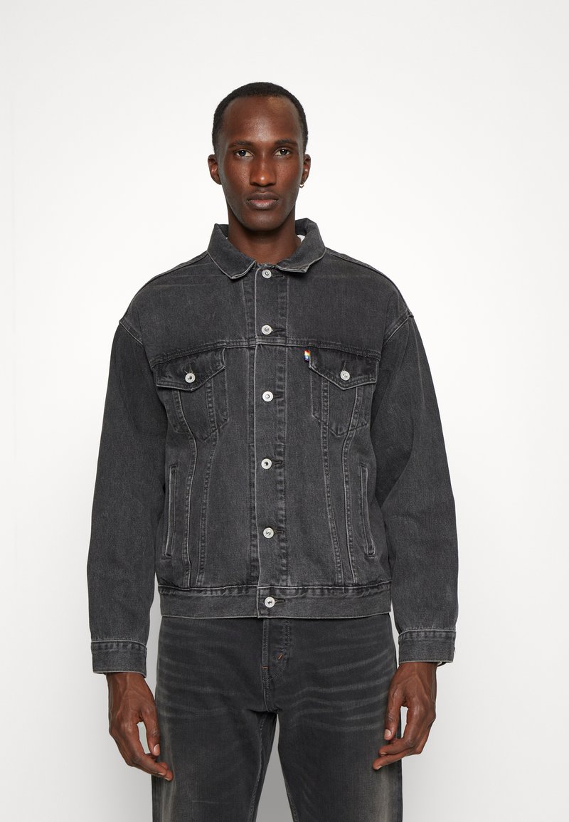 Chaqueta de mezclilla gris oscuro con cierre de botones, dos bolsillos en el pecho y un collar clásico. Presenta una textura ligeramente desgastada y un branding mínimo.