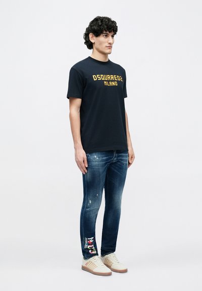 Mørkeblå bomuld T-shirt med gul "DSQUARED2 MILANO"-logo, kombineret med slidte denimjeans og beige sneakers med teksturerede detaljer.
