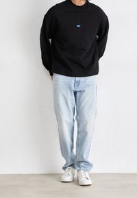 Hombre vestido con una camiseta de manga larga negra con un logo de Hugo, jeans ligeros y zapatillas blancas, de pie contra una pared blanca lisa.