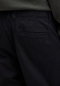 Gros plan sur l'arrière d'un pantalon noir avec poches passepoilées et logo brodé discret, associé à un haut gris.