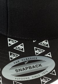 Musthave musta snapback-kate, millel on tekstureeritud kangas, valge kolmnurkade muster ääres ja hõbedane ovaalne logo kleebis koos tekstiga.