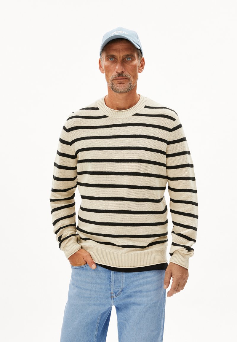 Pull rayé crème et noir, avec un col rond et des poignets côtelés, associé à un jean bleu clair et une casquette bleu clair.