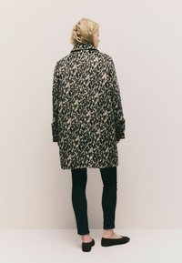 Blond osoba čelí stene a má na sebe kabát s leopardím vzorom po kolená, čierne slim nohavice a čierne balerínky.