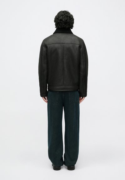 Juoda odinė striukė su kailio apykakle, turinti tiesų kirpimą ir dvi šonines kišenes, dėvima kartu su tamsiai žaliais platėjančiais kelnėmis.