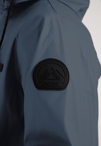 Chaqueta impermeable de tela azul oscuro. Presenta un parche de goma negra texturizada con un diseño de montaña y el logo "JACK1T" en la manga.