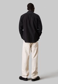 Camisa negra de manga larga con cuello, combinada con pantalones de pierna ancha en color crema y zapatos negros. Detalles de textura plana y ajuste relajado visibles.