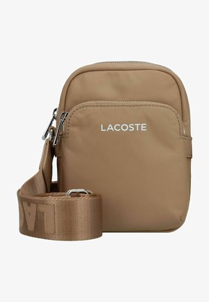 Sac bandoulière marron avec un extérieur en tissu lisse, une poche zippée à l'avant et une large sangle texturée portant le nom de la marque "LACOSTE."