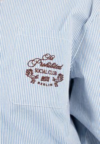 Blau-weiß gestreifte Hemd mit einer Brusttasche, verziert mit braunemgesticktem Text und Pferdemotiven von "The Prohibited Social Club, Berlin."